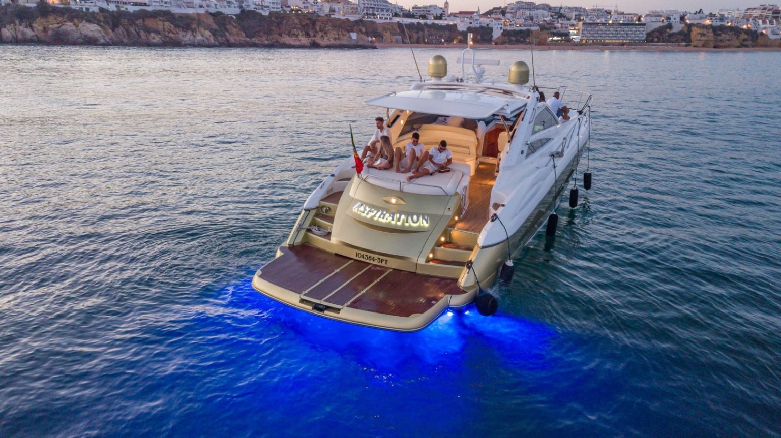 Iate Sunseeker privado ao entardecer, com luzes subaquáticas e a Baixa de Albufeira ao fundo